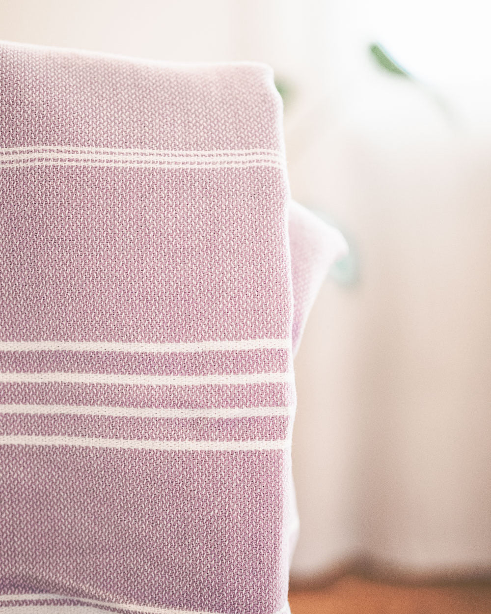 Eine Nahaufnahme des Maxi Strandtuchs De La Mer von Main Design, einer gefalteten lavendelfarbenen und weiß gestreiften Strickdecke aus 100 % Baumwolle, gezeigt mit weicher Beleuchtung und unscharfem Hintergrund.