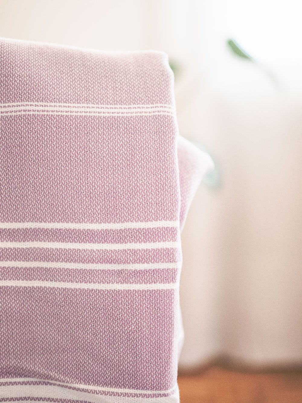 Eine Nahaufnahme des Maxi Strandtuchs De La Mer von Main Design, einer gefalteten lavendelfarbenen und weiß gestreiften Strickdecke aus 100 % Baumwolle, gezeigt mit weicher Beleuchtung und unscharfem Hintergrund.