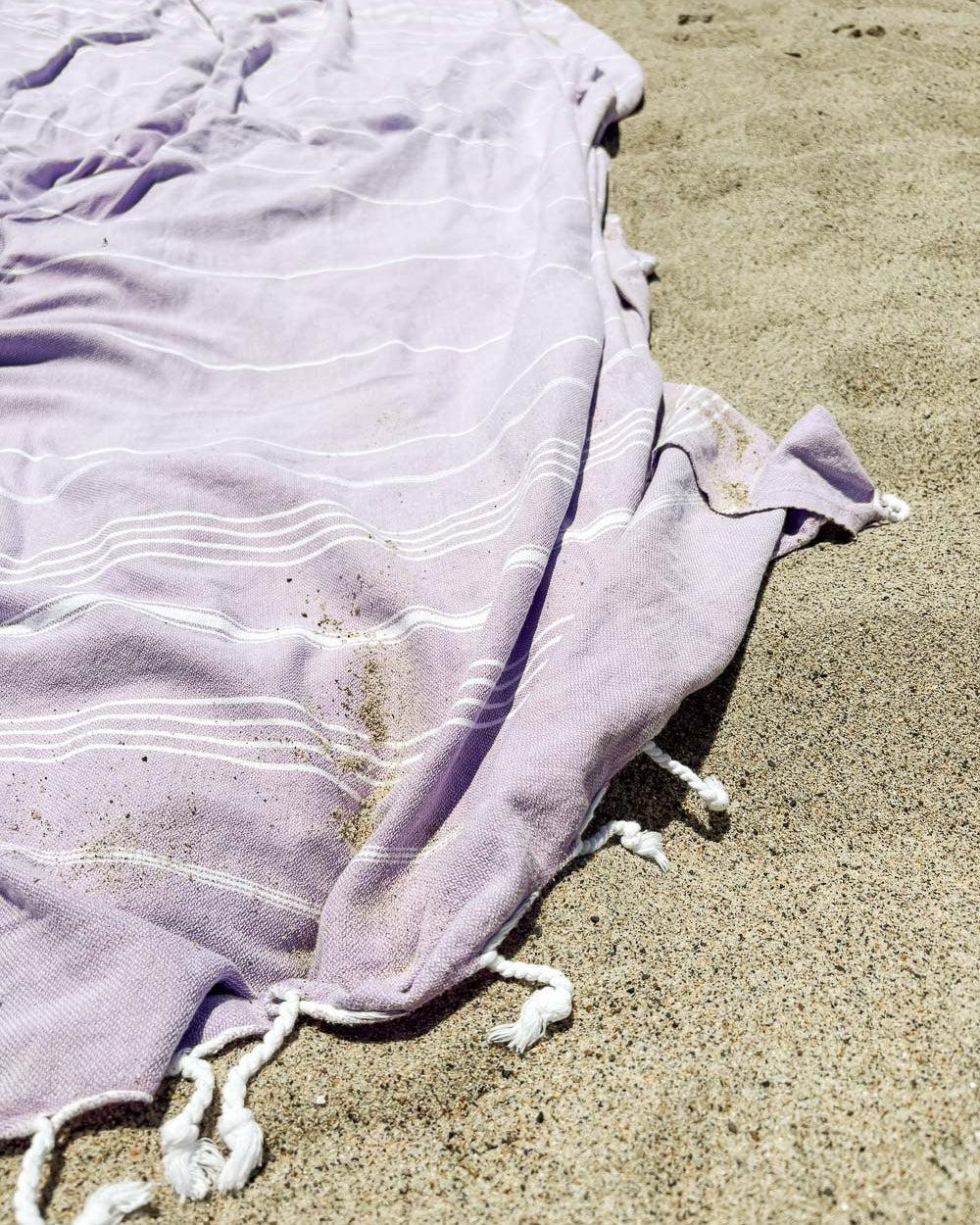 Das lilafarbene Maxi Strandtuch De La Mer von Main Design aus 100% Baumwolle mit weißen Streifen und geknoteten Fransen liegt auf dem Sand, etwas Sand bedeckt das OEKO-TEX-zertifizierte Strandtuch.