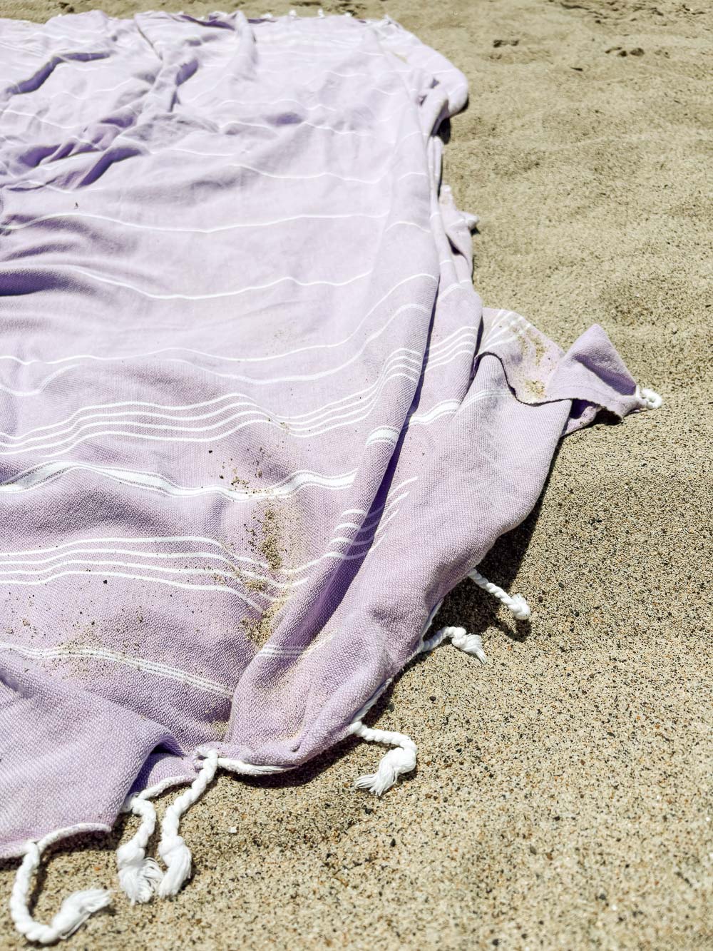 Das lilafarbene Maxi Strandtuch De La Mer von Main Design aus 100% Baumwolle mit weißen Streifen und geknoteten Fransen liegt auf dem Sand, etwas Sand bedeckt das OEKO-TEX-zertifizierte Strandtuch.