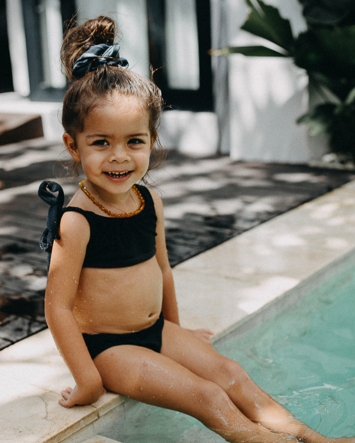 Ein lächelndes kleines Kind sitzt am Rand eines Pools und lässt seine Füße im Wasser baumeln. Das Kind trägt einen schwarzen Mia Mädchen Bikini von Main Design mit LSF 50+ und hat seine Haare zu einem Knoten mit einer Schleife zusammengebunden. Dahinter sind einige Pflanzen und die Außenseite eines Gebäudes ist sichtbar.