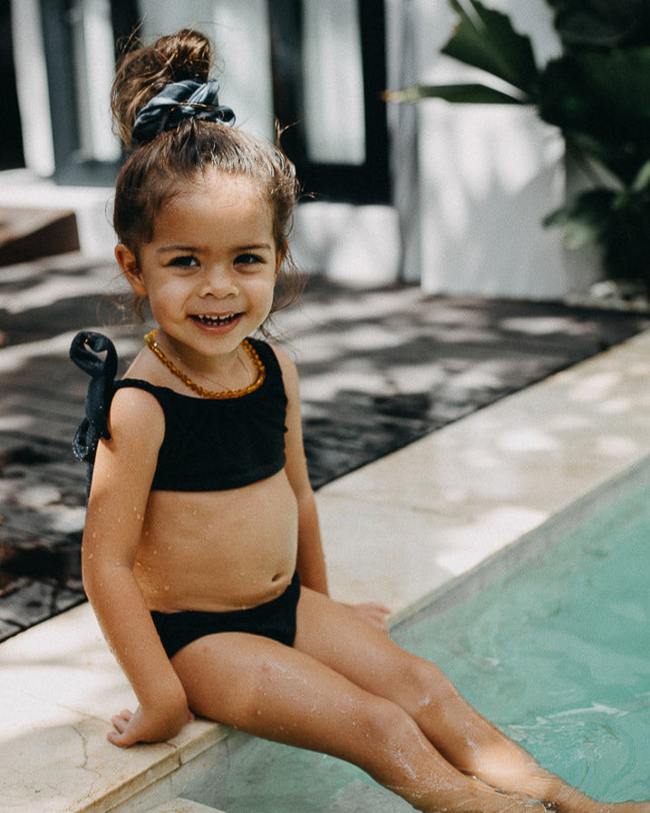 Ein lächelndes kleines Kind sitzt am Rand eines Pools und lässt seine Füße im Wasser baumeln. Das Kind trägt einen schwarzen Mia Mädchen Bikini von Main Design mit LSF 50+ und hat seine Haare zu einem Knoten mit einer Schleife zusammengebunden. Dahinter sind einige Pflanzen und die Außenseite eines Gebäudes ist sichtbar.