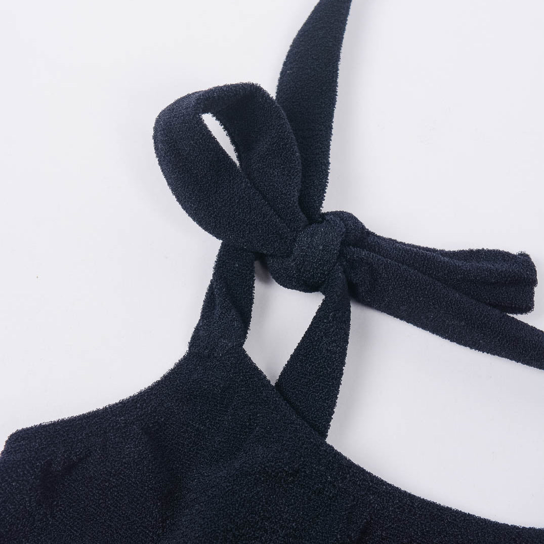 Eine Nahaufnahme des Main Design Mia Mädchen Bikinis, aus dunkelmarineblauem Stoff mit strukturierter Oberfläche. Das Bikinioberteil aus recycelten Materialien zeigt sein elegantes Neckholder-Design mit einem ordentlich gebundenen Knoten an einer Ecke. Der schlichte weiße Hintergrund hebt die komplizierten Details dieses Kleidungsstücks hervor.