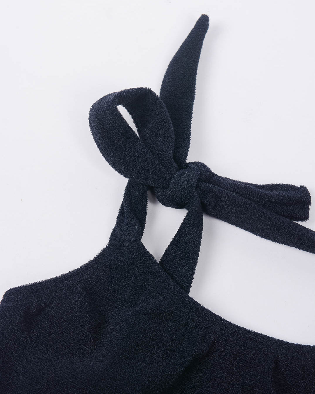 Eine Nahaufnahme des Main Design Mia Mädchen Bikinis, aus dunkelmarineblauem Stoff mit strukturierter Oberfläche. Das Bikinioberteil aus recycelten Materialien zeigt sein elegantes Neckholder-Design mit einem ordentlich gebundenen Knoten an einer Ecke. Der schlichte weiße Hintergrund hebt die komplizierten Details dieses Kleidungsstücks hervor.