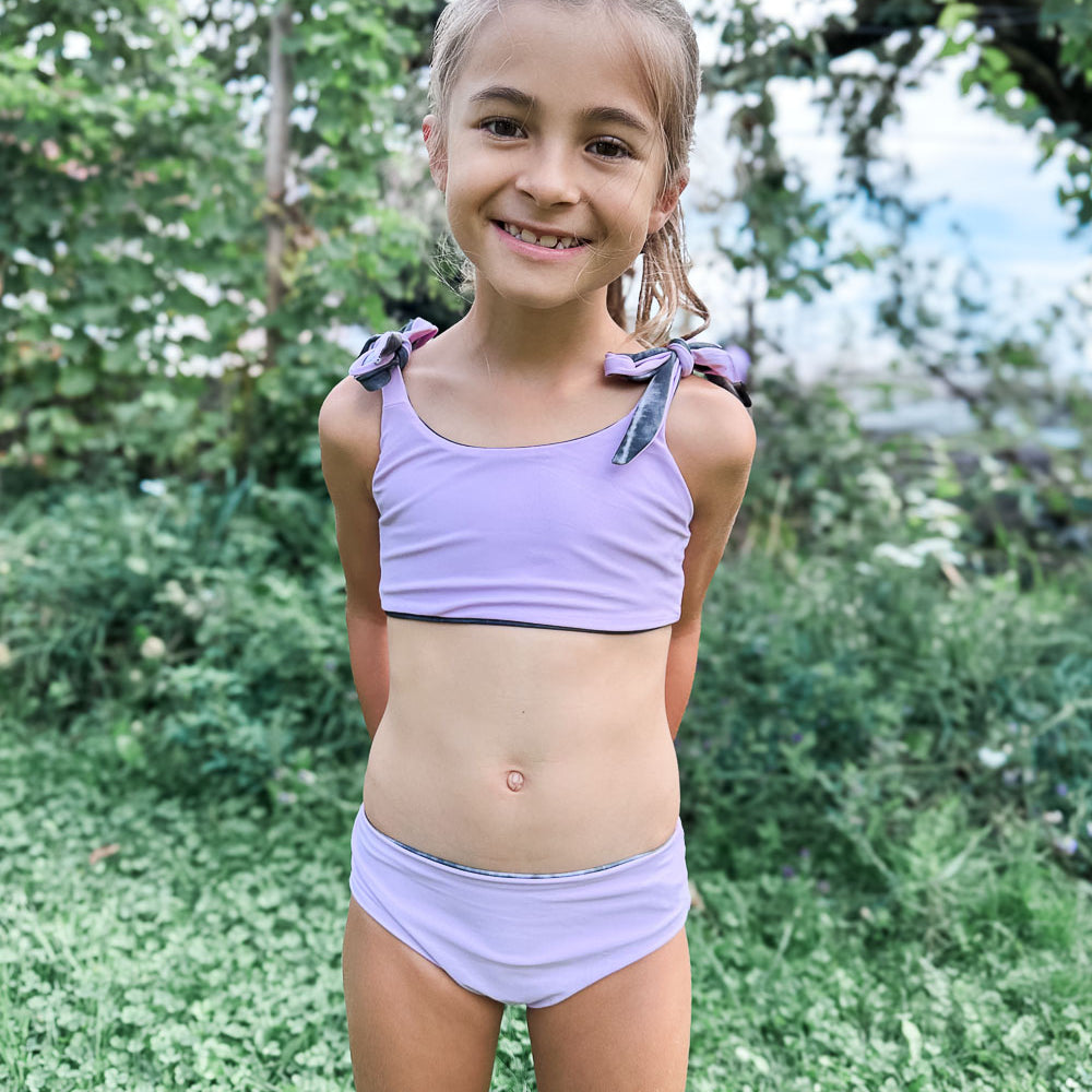 Ein junges Mädchen lächelt in die Kamera. Es steht auf grünem Gras und trägt den Mia Mädchen Bikini reversibel von Main Design - einen hellvioletten, nachhaltigen Bikini mit Schleifendetails. Im Hintergrund sind Bäume und Pflanzen zu sehen.