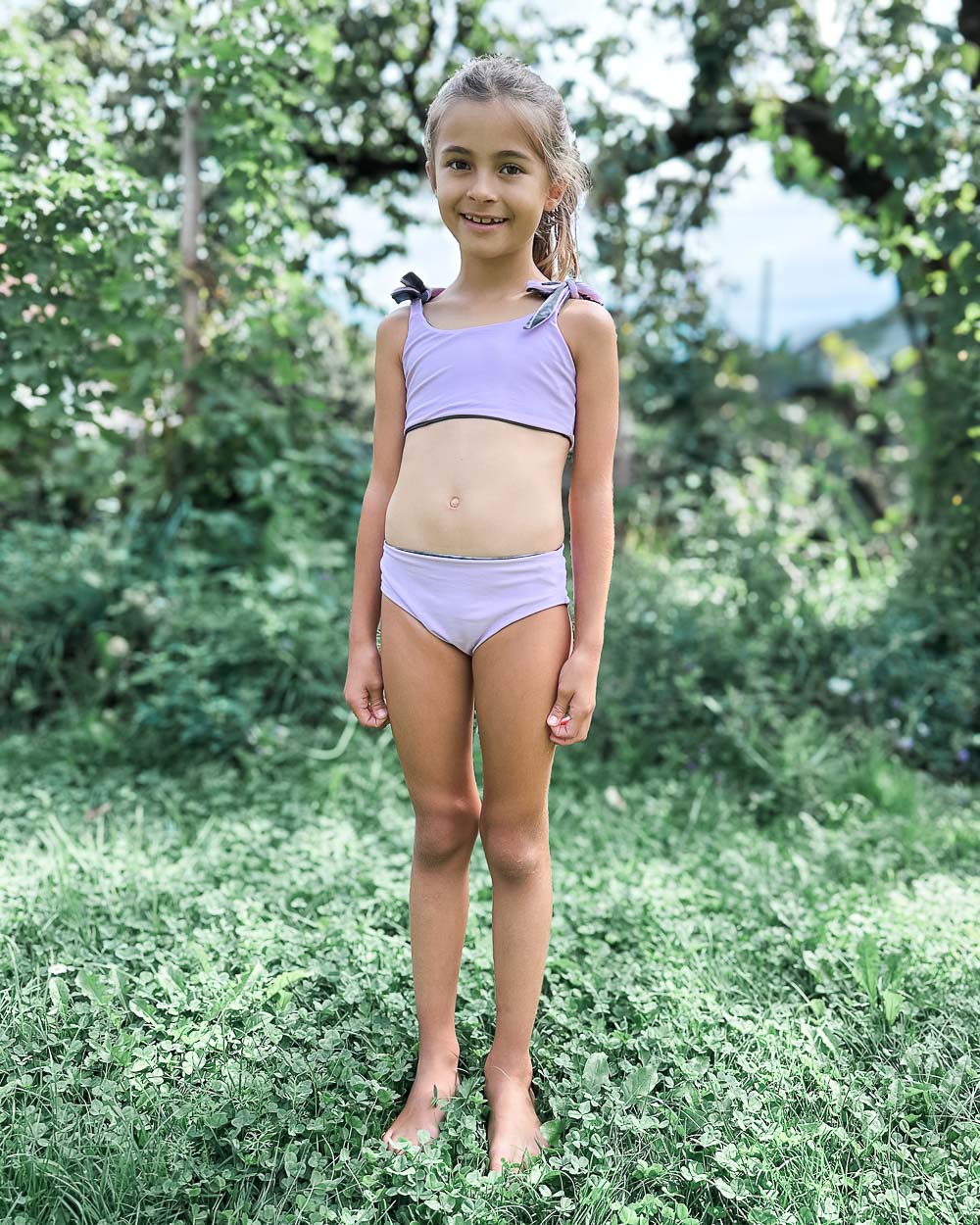 Ein junges Mädchen mit hellbraunen Zöpfen steht barfuß im Gras und lächelt in dem Main Design Mia Mädchen Bikini reversibel, einem hellvioletten zweiteiligen Badeanzug. Sie ist im Freien, umgeben von grünen Bäumen und Pflanzen.