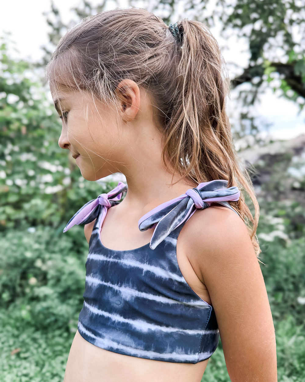 Junges Mädchen mit langen braunen Haaren in einem Pferdeschwanz trägt den Mia Mädchen Bikini reversibel von Main Design, mit lilafarbenen Schulterbändern. Sie steht im Freien vor einer Grünfläche, schaut nach links und lächelt sanft.