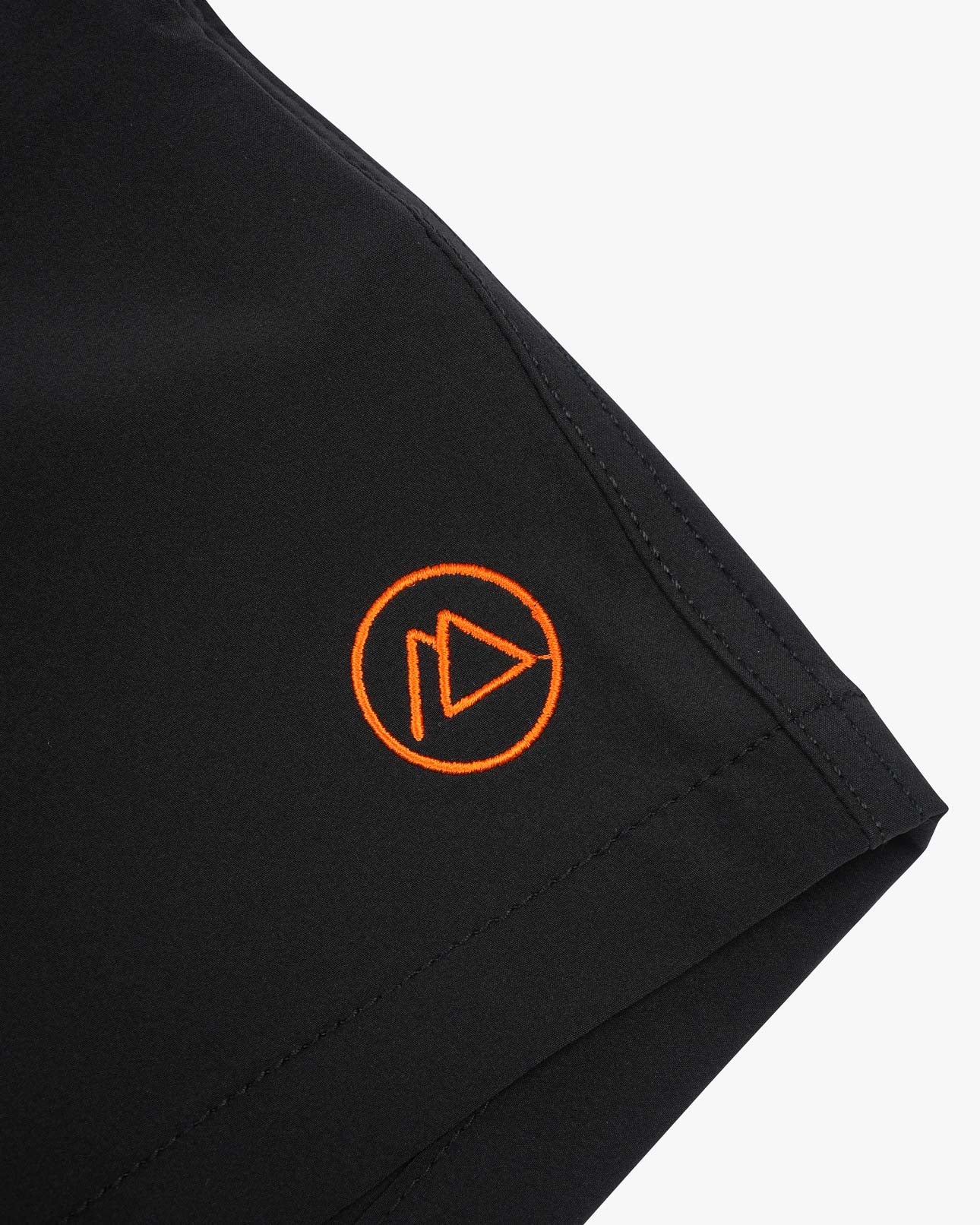 Nahaufnahme der schwarzen Main Design Nolan Boardshorts für Jungen mit einem kleinen orangefarbenen, kreisförmigen Logo mit Dreieck und geschwungener Linie. Der glatte Stoff bietet LSF 50+ Sonnenschutz, mit sichtbaren Nähten entlang der Naht, die das 4-Wege-Stretch-Material-Design betonen.