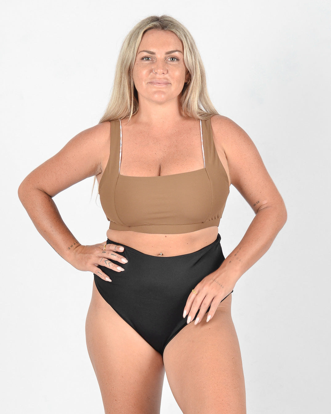Eine Frau mit langen blonden Haaren steht vor einem schlichten weißen Hintergrund. Sie trägt die Posh Bikinihose von Main Design mit einem braunen Oberteil und einer hoch taillierten schwarzen Hose aus umweltfreundlichem Stoff. Ihre Arme hängen entspannt an ihren Seiten und sie lächelt leicht in die Kamera.