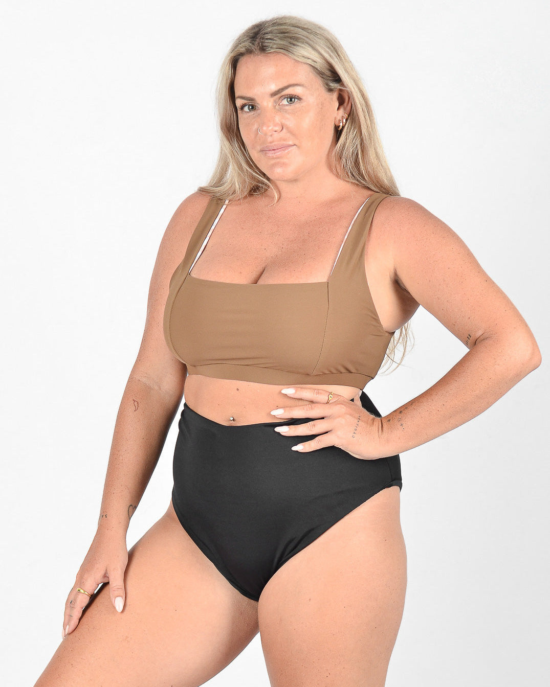 Eine Frau mit langen blonden Haaren steht vor einem schlichten weißen Hintergrund. Sie trägt ein braunes bauchfreies Top aus umweltfreundlichem Stoff und eine hoch taillierte schwarze Hose von Main Design namens Posh Bikini Hose. Ihre rechte Hand liegt auf ihrer Hüfte und ihr linker Arm ruht an ihrer Seite. Sie hat einen selbstbewussten Ausdruck und mehrere Tattoos auf ihren Armen und Fingern.
