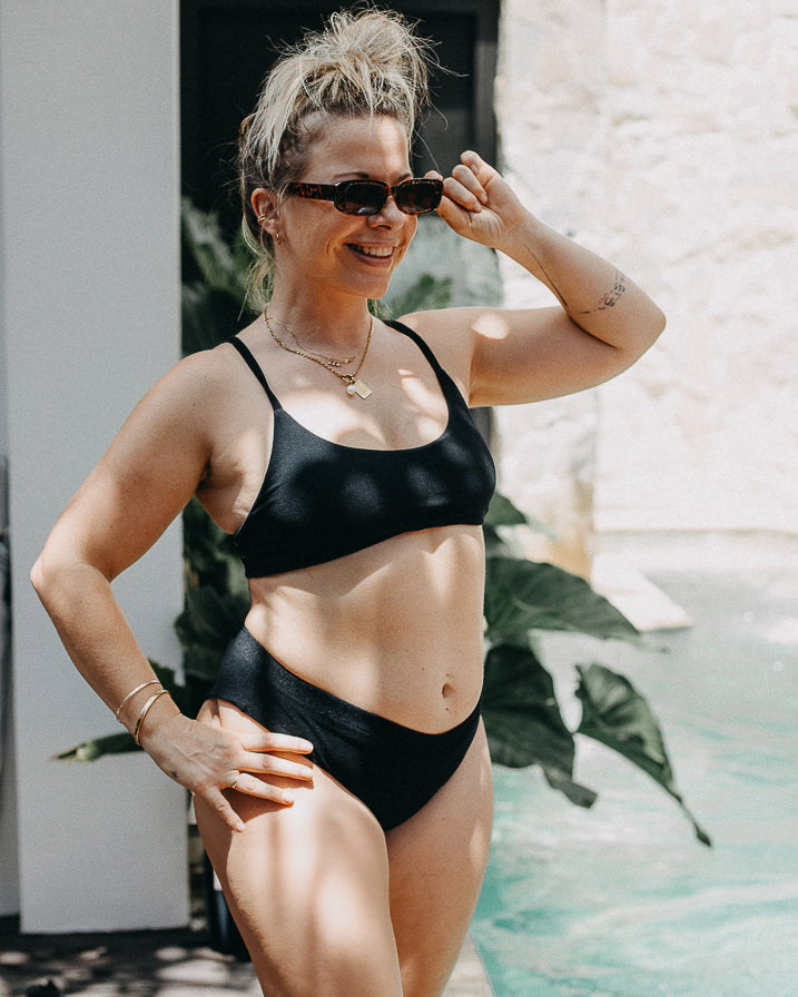 Eine Frau in einem Main Design Sassy Bikini-Top und Sonnenbrille steht lächelnd am Pool, die Haare hochgesteckt und die Sonnenbrille in der Hand. Sonnenlicht und grüne Pflanzen bilden einen lebendigen Hintergrund.