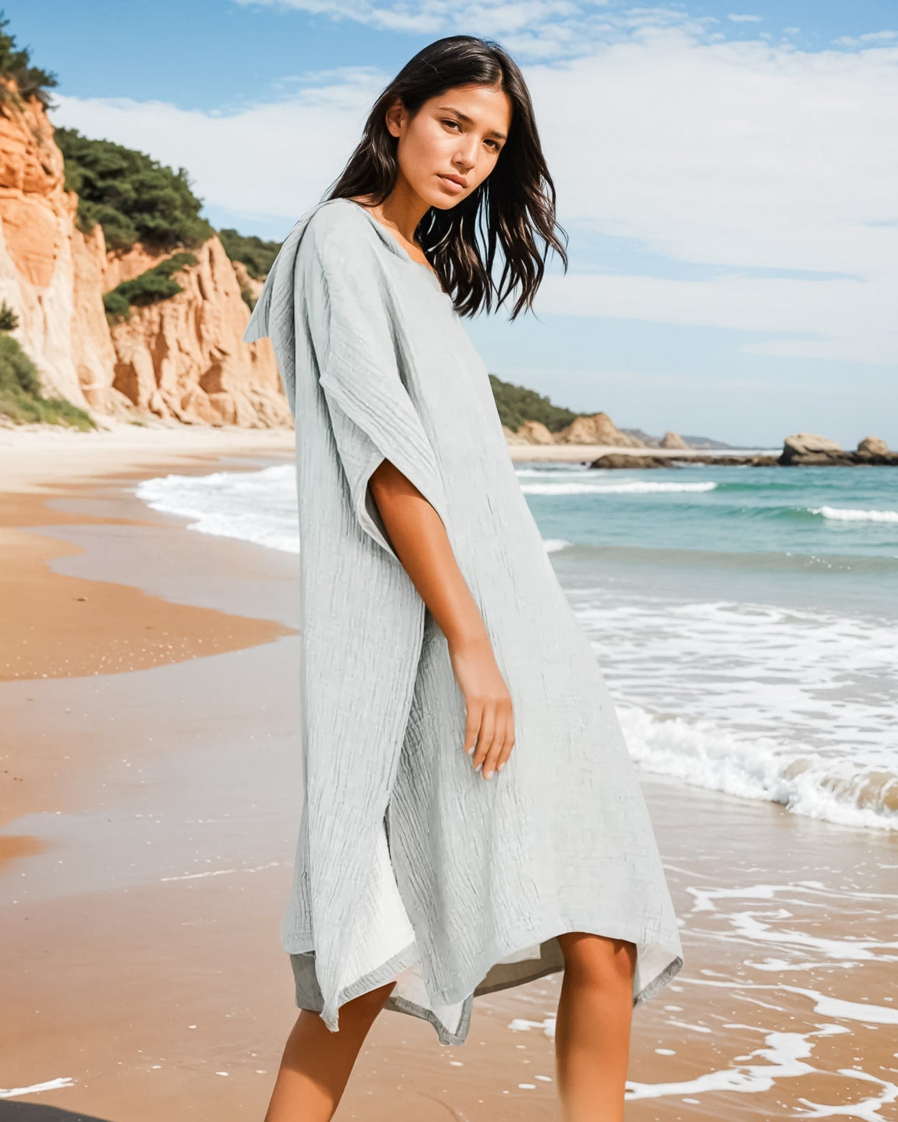 Eine Frau im Main Design Shoreline Breeze Poncho, einem lockeren hellgrauen Kleid, steht an einem Sandstrand mit Klippen und Meer im Hintergrund unter einem teilweise bewölkten Himmel - ideal für entspannte Strandtage.