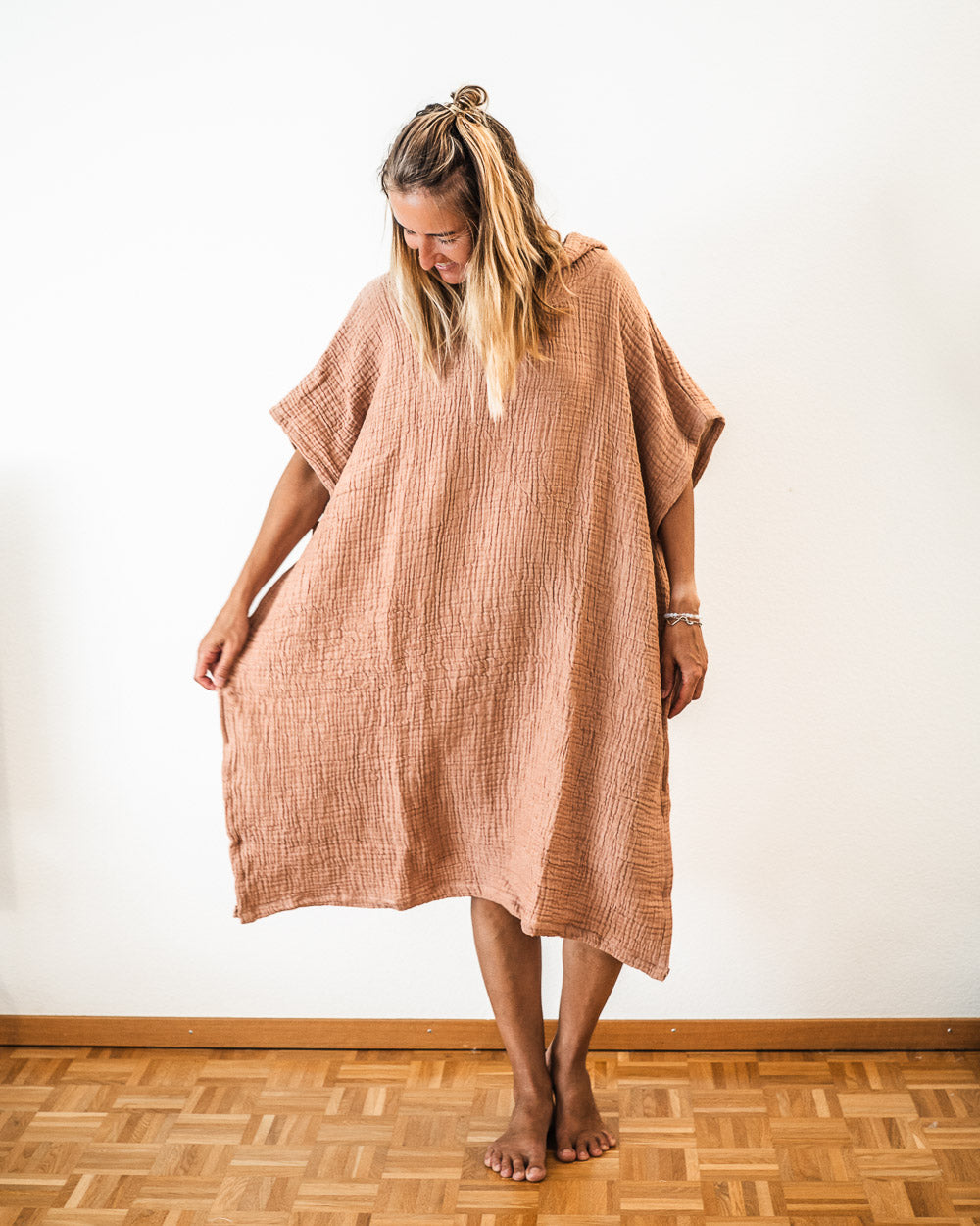 Eine Frau mit langen blonden Haaren steht barfuß auf einem Holzboden, lächelt und schaut nach unten, während sie einen lockeren, strukturierten, hellbraunen Shoreline Breeze Poncho von Main Design in Einheitsgröße trägt. Sie hält die Seiten des Ponchos sanft nach außen.
