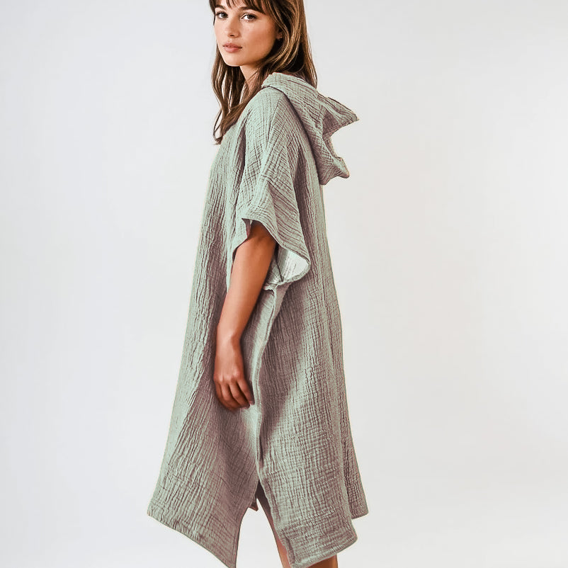 Eine Frau steht seitlich in dem Main Design Shoreline Breeze Poncho, einem lockeren, strukturierten hellgrauen Kleidungsstück mit kurzen Ärmeln und knielangem Saum in Einheitsgröße, das vor einem schlichten weißen Hintergrund gestylt ist.