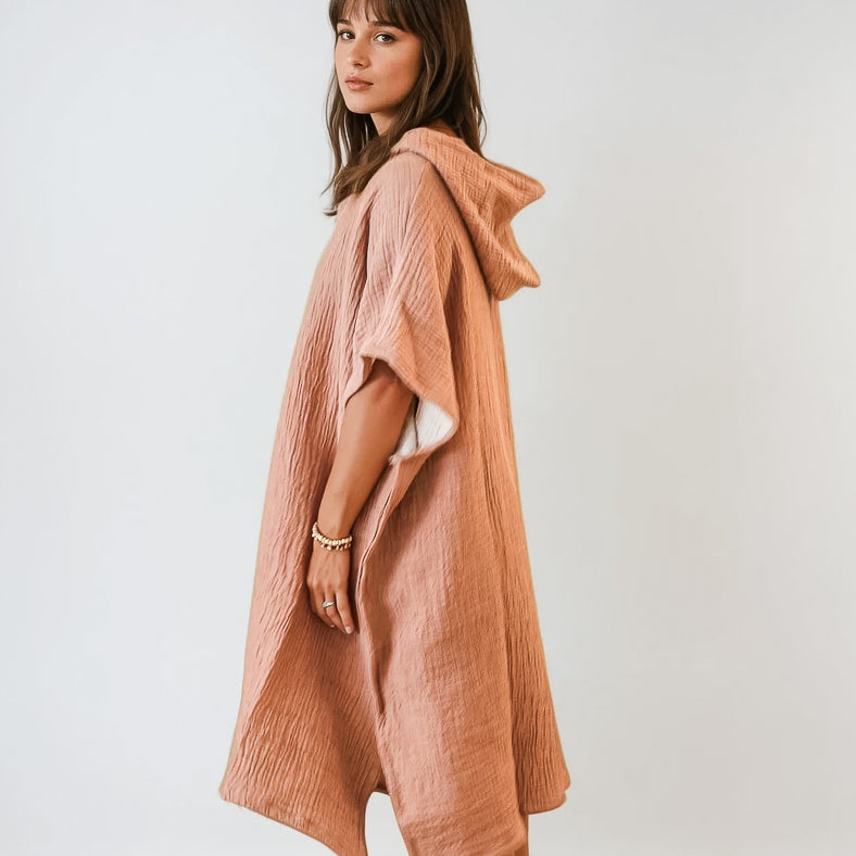 Eine Frau steht seitlich und trägt einen lockeren, strukturierten, hellbraunen Shoreline Breeze Poncho von Main Design mit kurzen Ärmeln. Sie hat mittellanges braunes Haar und blickt in die Kamera vor einem schlichten weißen Hintergrund. Eine Größe passt den meisten.