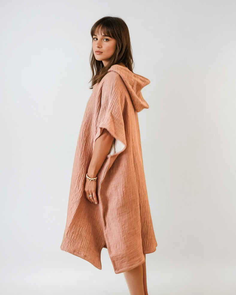 Eine Frau steht seitlich und trägt einen lockeren, strukturierten, hellbraunen Shoreline Breeze Poncho von Main Design mit kurzen Ärmeln. Sie hat mittellanges braunes Haar und blickt in die Kamera vor einem schlichten weißen Hintergrund. Eine Größe passt den meisten.