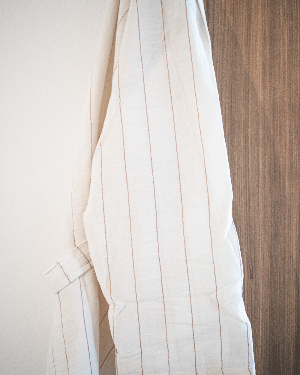 Der Kimono Soleil von Main Design aus 100% Baumwolle mit cremefarbenem Streifenmuster hängt auf einer Holzfläche mit sanftem natürlichem Licht.