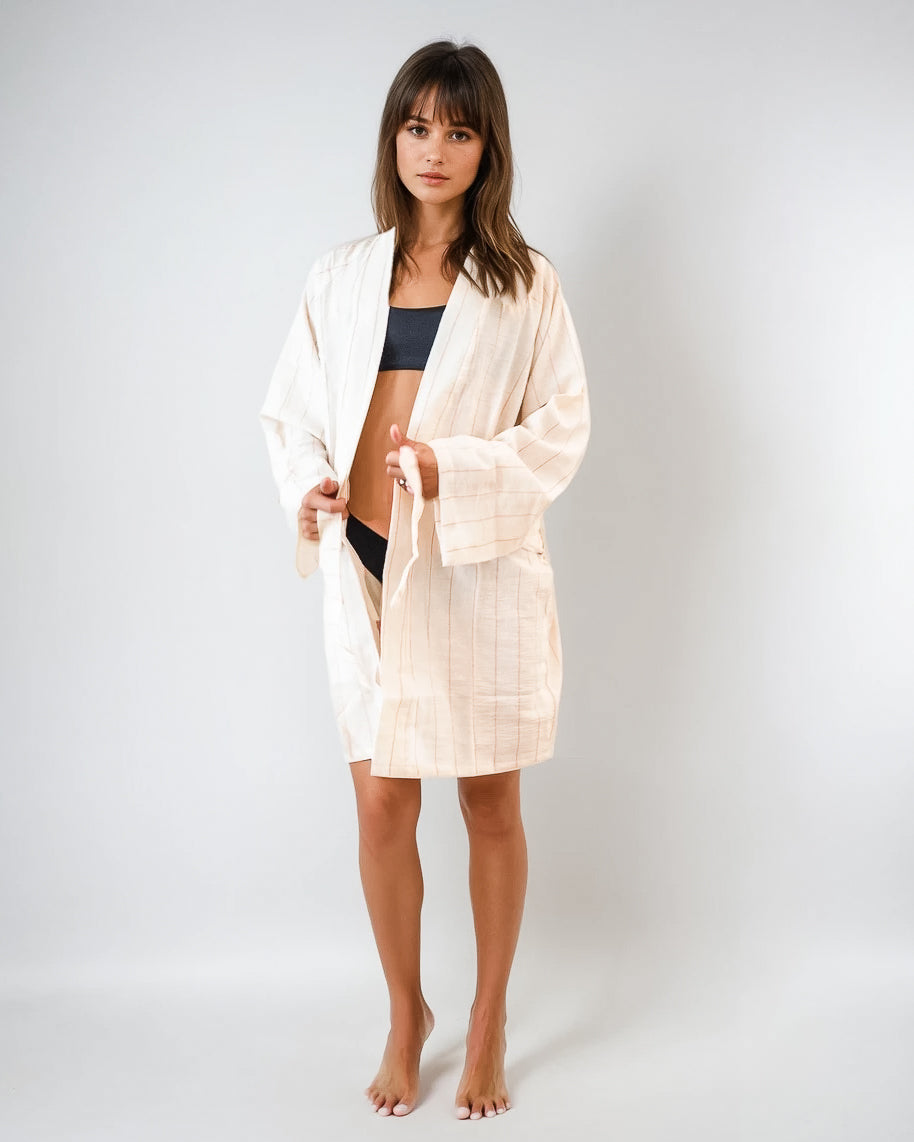 Eine Frau steht barfuß vor einem schlichten Hintergrund, trägt den Main Design Soleil Kimono - einen hellen, lockeren Bademantel aus 100 % Baumwolle - über einem schwarzen Bikini oder Unterwäsche und blickt mit neutralem Blick direkt in die Kamera.