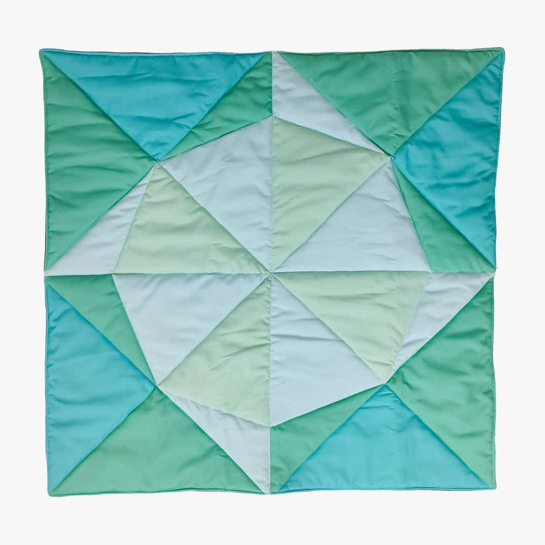 Eine lebendige, handgefertigte Spielmatte und Krabbeldecke Boot von Main Design zeigt ein geometrisches Muster mit unterschiedlichen Grün-, Türkis- und hellblauen Dreiecken auf weißem Hintergrund. Ideal als schicke Krabbelmatte für Ihren Kleinen.