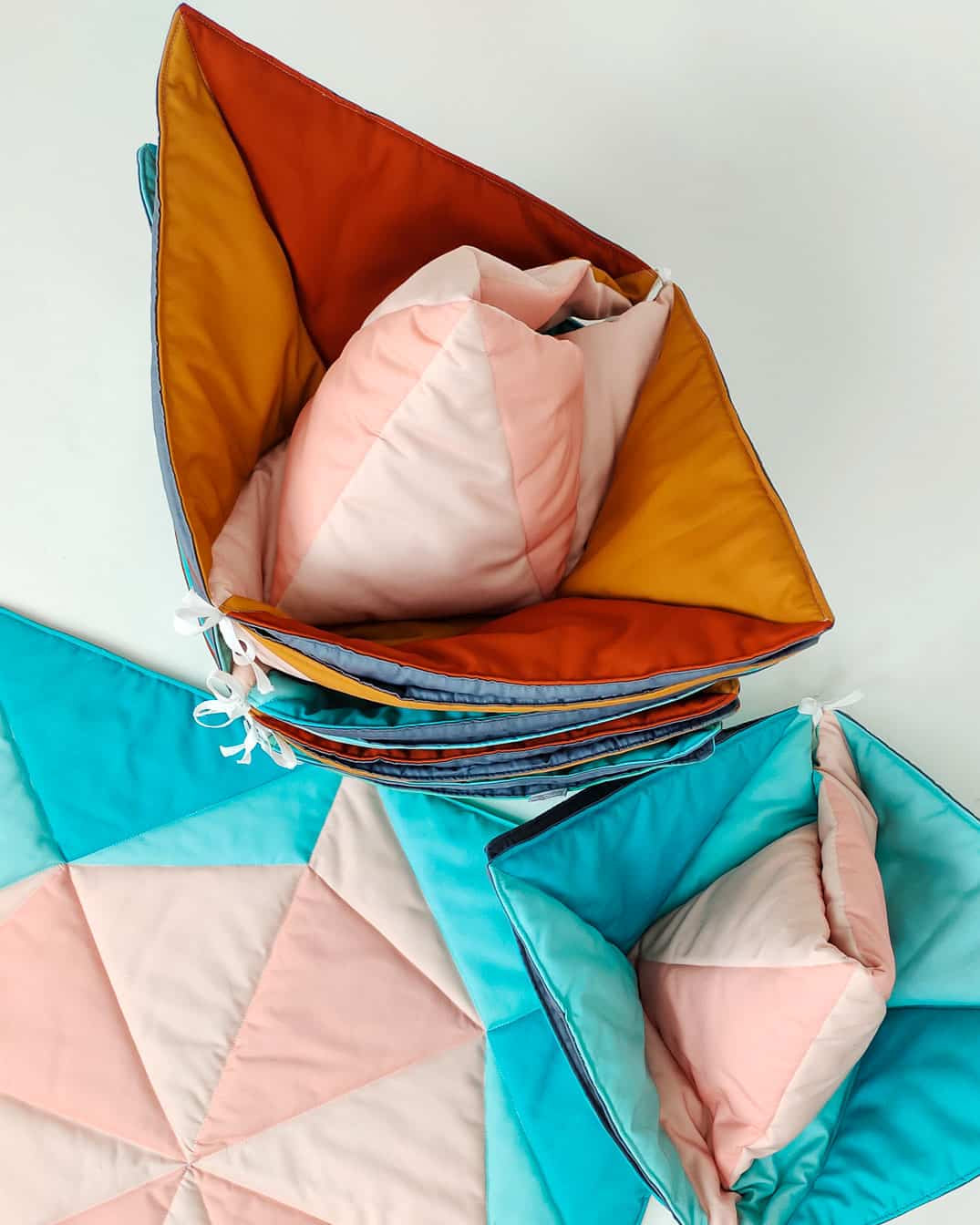 Ein Set aus farbenfrohen, geometrischen Steppkörben und einer dazu passenden handgefertigten Spielmatte und Krabbeldecke Boot von Main Design. Die weichen Körbe zeigen ein blaues, orangefarbenes und rosafarbenes Dreiecksmuster, während die Matte mit einem rosafarbenen und blauen Sternenmuster verziert ist. Diese Körbe lassen sich bequem ineinander stapeln und so leicht verstauen.