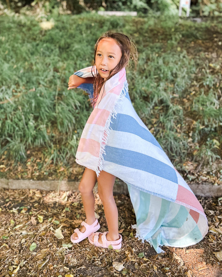 Ein junges Mädchen steht im Freien auf grünem Grund, eingewickelt in das Baumwollhandtuch "Strandtuch Blockberry" von Main Design. Sie trägt rosa Sandalen und schaut mit nassen Haaren zur Seite, umgeben von Gras und Bäumen.