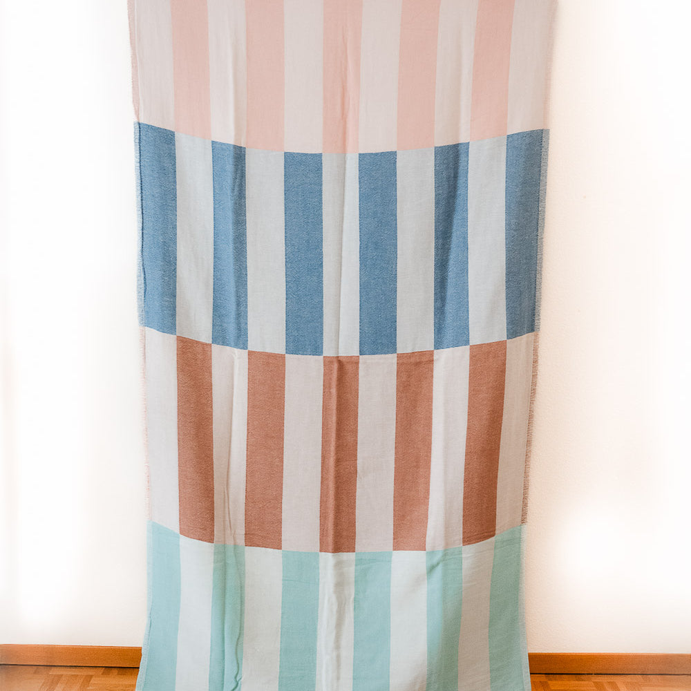 Das Strandtuch Blockberry von Main Design ist rechteckig, mit kurzen Fransen und vertikalen Pastellstreifen in Pink, Blau, Braun und Mintgrün - erinnert an ein buntes Strandtuch, flach an einer weißen Wand über Holzboden hängend.