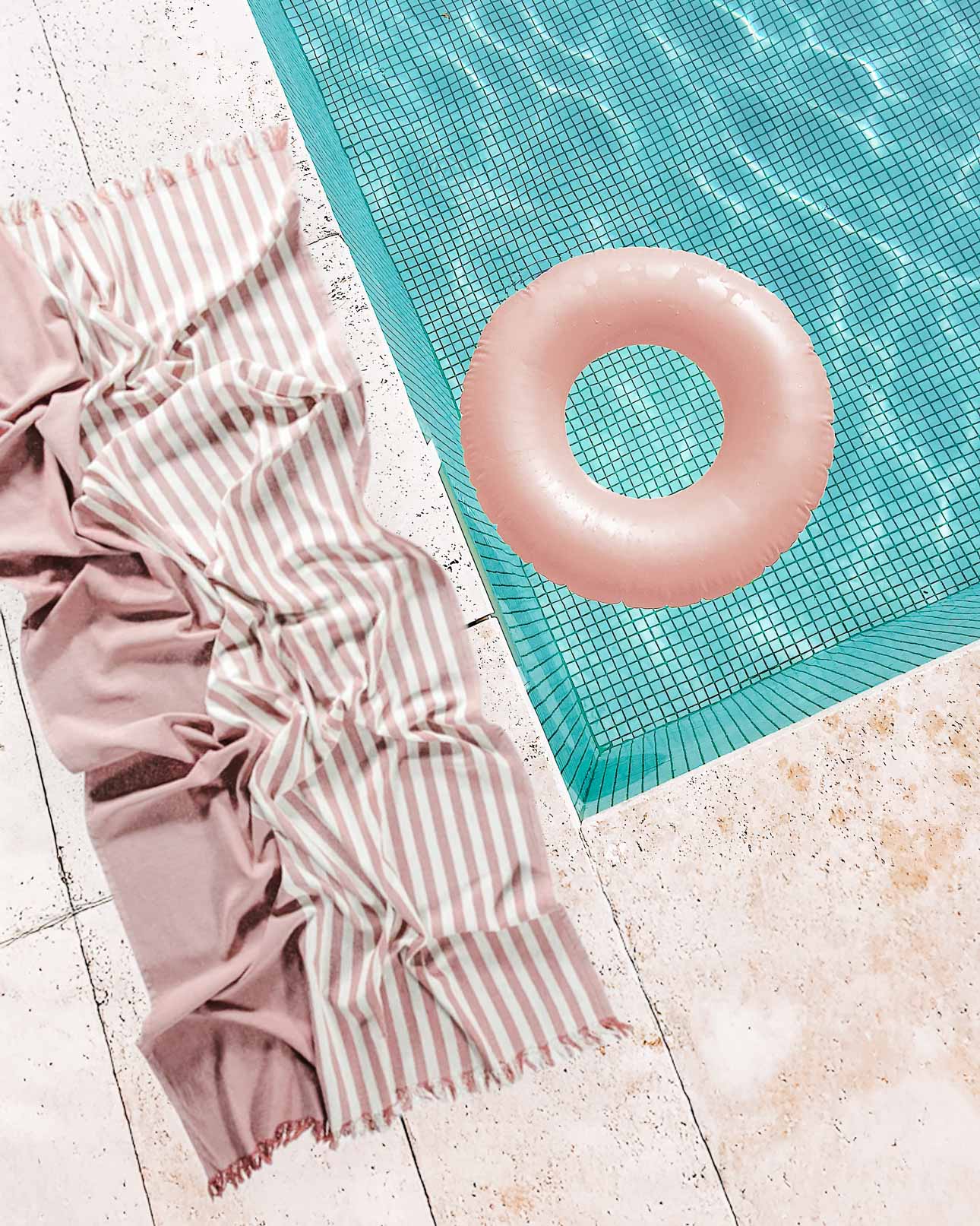 Ein rosa-weiß gestreiftes Main Design Strandtuch Fiesta liegt zerknittert auf dem gefliesten Beckenrand neben einem hellrosa aufblasbaren Ring, der im blauen Becken schwimmt.