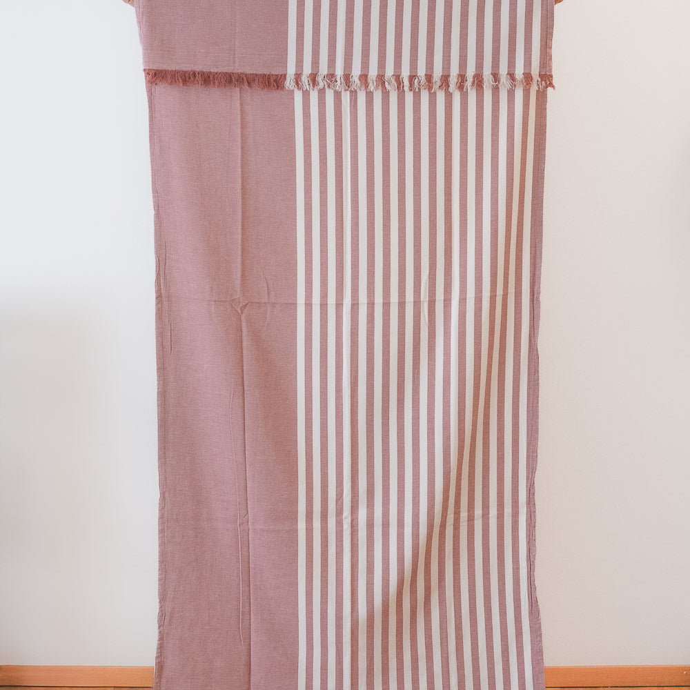 Das Main Design Strandtuch Fiesta aus OEKO-TEX-Baumwolle zeigt vertikale cremefarbene und gedeckte rosa Streifen auf der einen Seite und einfarbiges gedecktes Rosa auf der anderen Seite, die vor einer weißen Wand und einem Holzboden hängen.