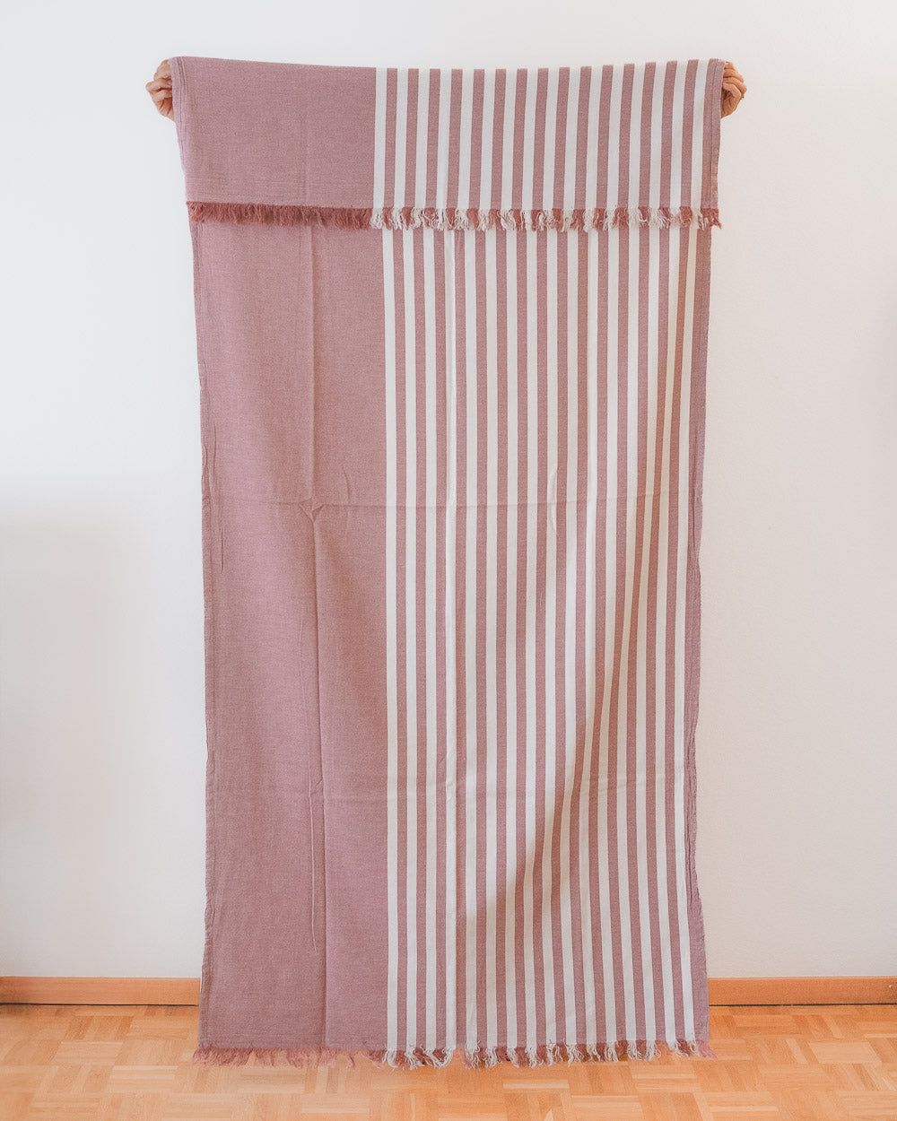 Das Main Design Strandtuch Fiesta aus OEKO-TEX-Baumwolle zeigt vertikale cremefarbene und gedeckte rosa Streifen auf der einen Seite und einfarbiges gedecktes Rosa auf der anderen Seite, die vor einer weißen Wand und einem Holzboden hängen.