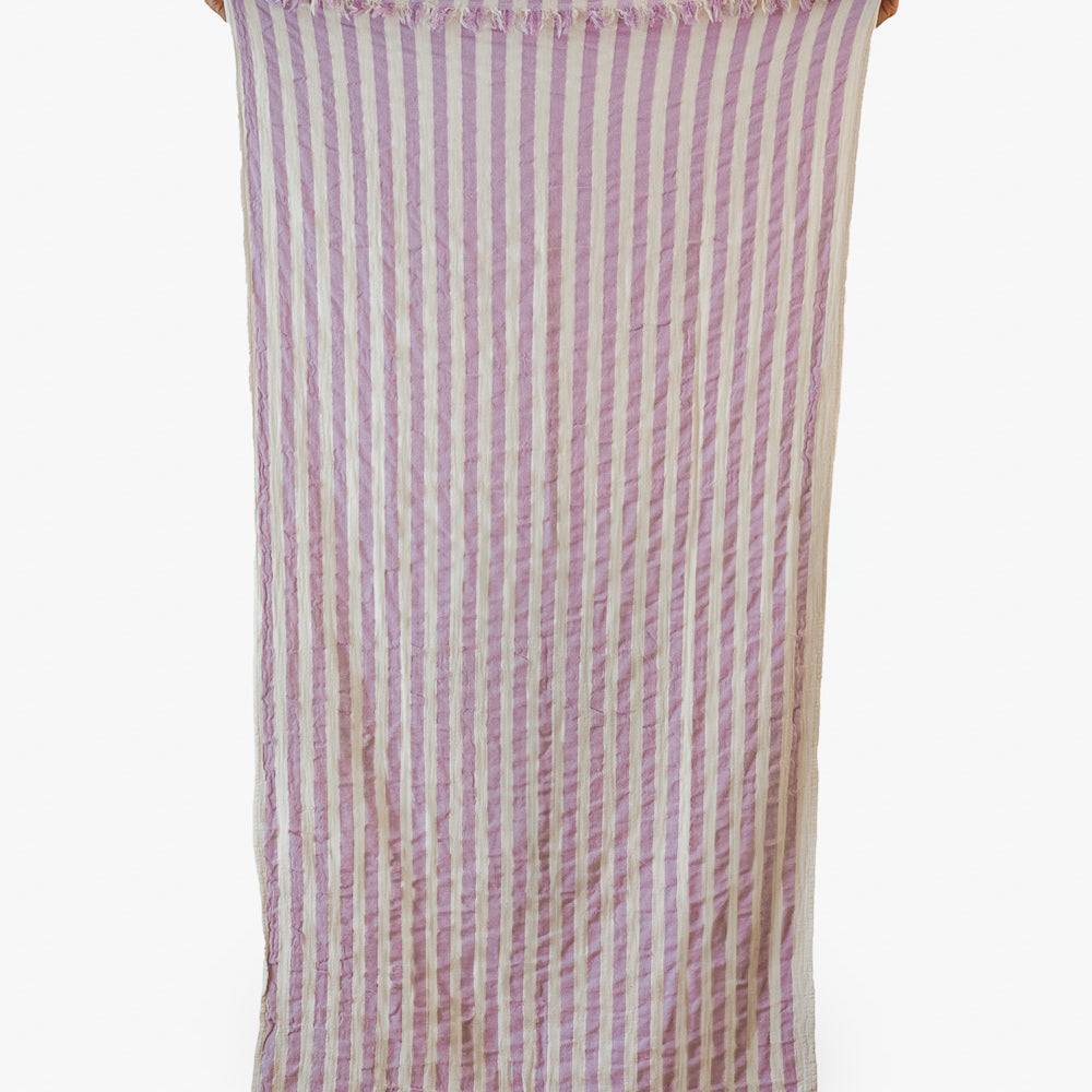 Das Strandtuch Melody von Main Design aus recycelter Baumwolle mit lavendel-weißen Streifen, kleinen Fransen und leichter Knitteroptik hängt vor schlicht weißem Hintergrund.