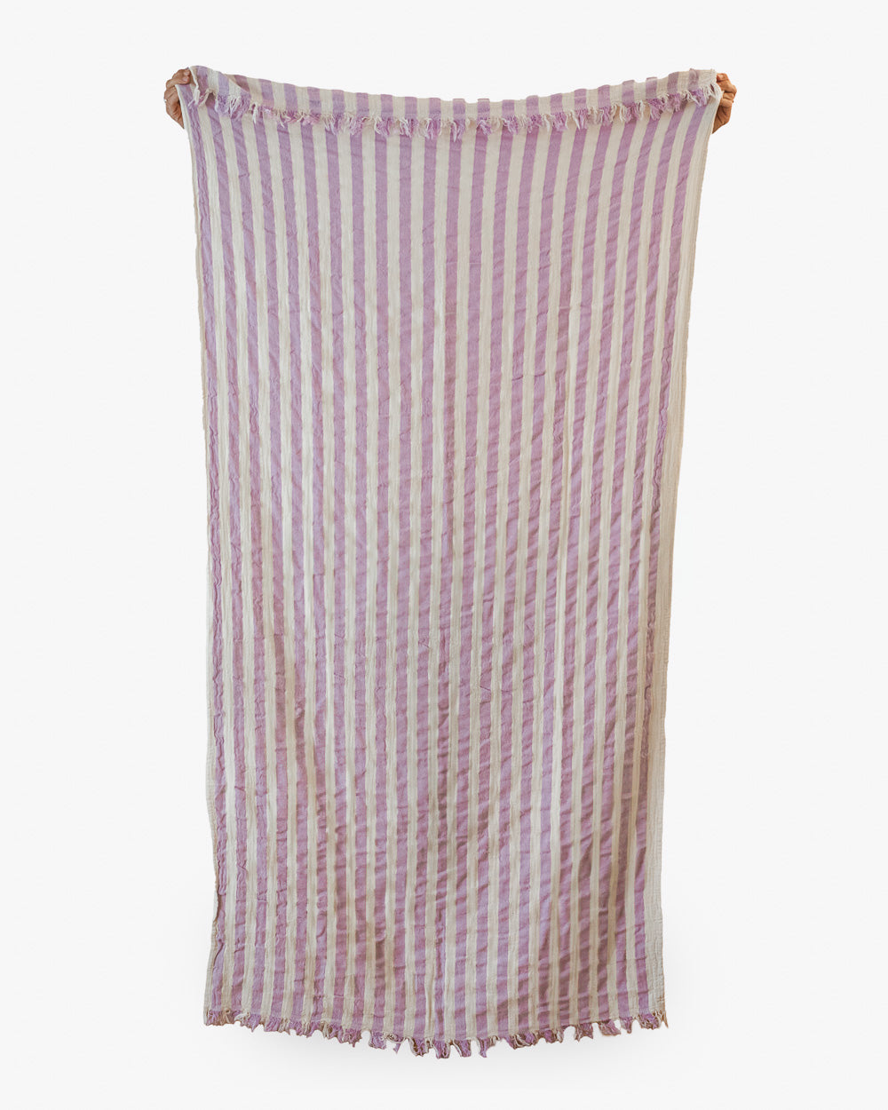 Das Strandtuch Melody von Main Design aus recycelter Baumwolle mit lavendel-weißen Streifen, kleinen Fransen und leichter Knitteroptik hängt vor schlicht weißem Hintergrund.