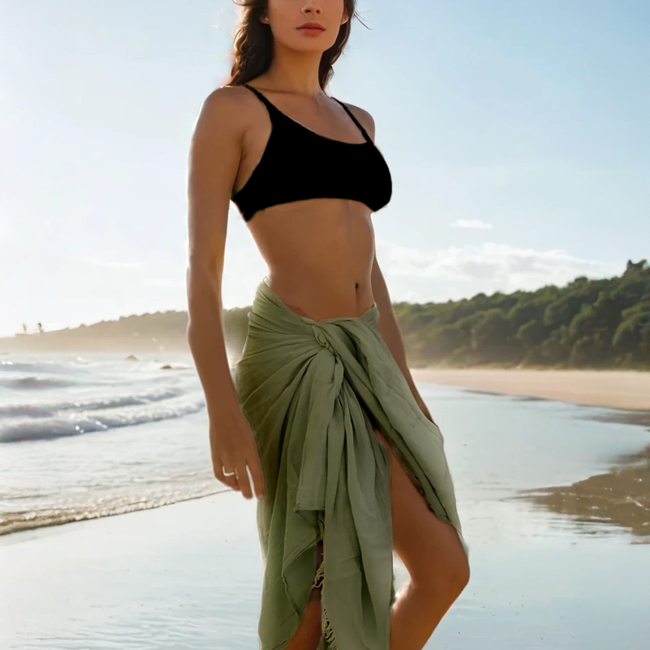 Bei Sonnenuntergang an einem Sandstrand hält eine Frau in einem schwarzen Crop-Top und einem hellgrünen Wickelrock das Strandtuch Nomad von Main Design aus OEKO-TEX-zertifizierter Baumwolle. Sanfte Wellen rollen heran und Bäume säumen das ferne Ufer.