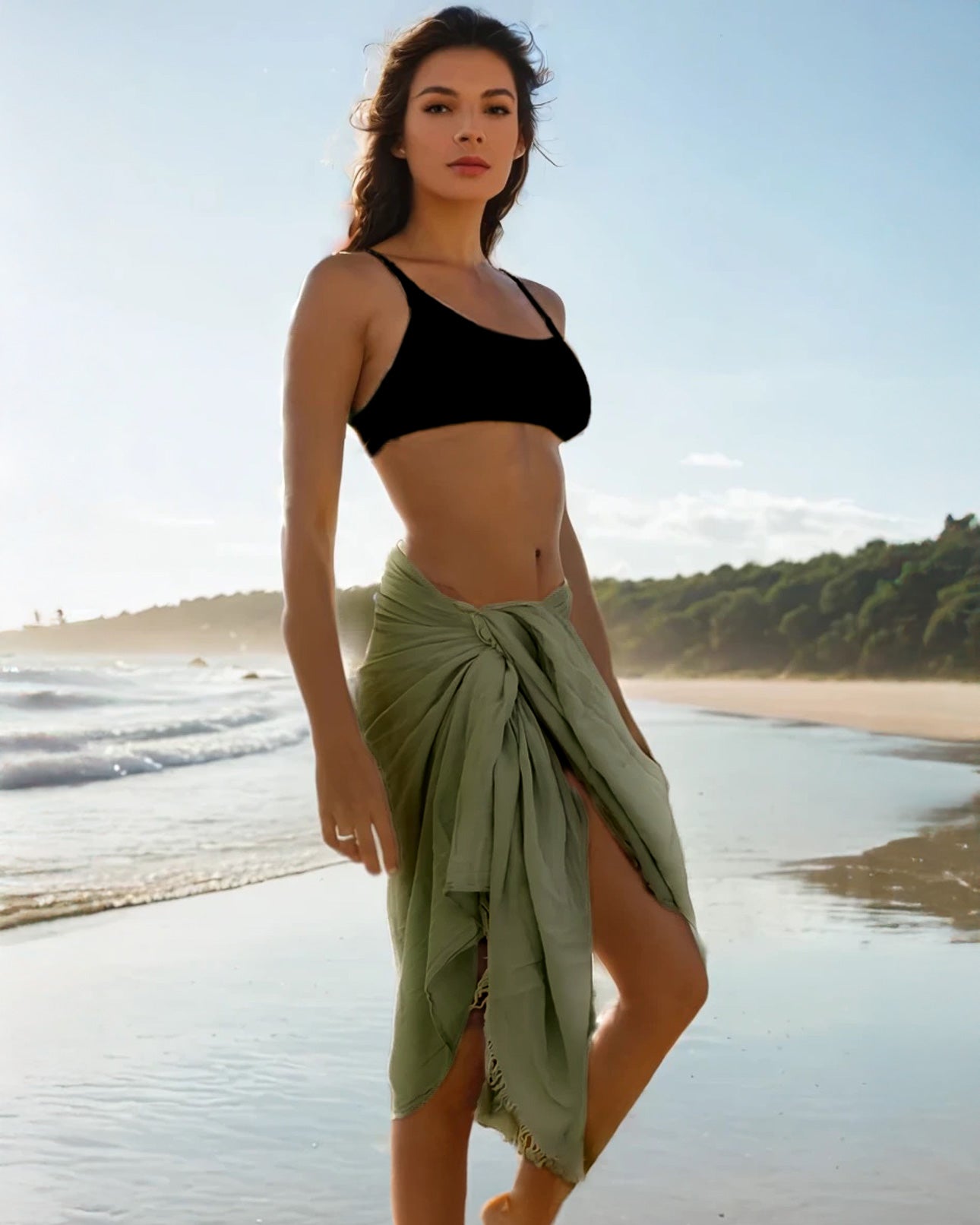 Bei Sonnenuntergang an einem Sandstrand hält eine Frau in einem schwarzen Crop-Top und einem hellgrünen Wickelrock das Strandtuch Nomad von Main Design aus OEKO-TEX-zertifizierter Baumwolle. Sanfte Wellen rollen heran und Bäume säumen das ferne Ufer.