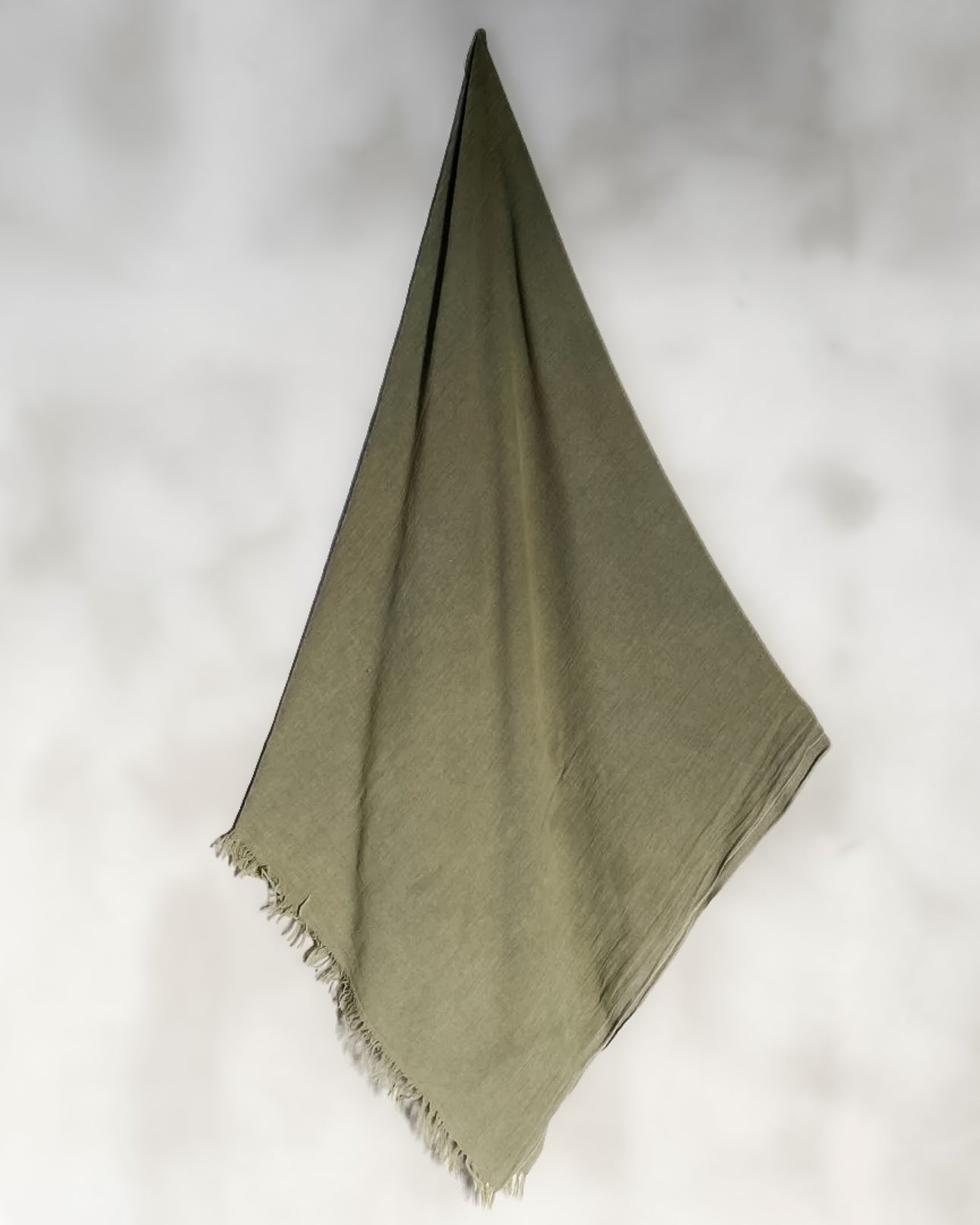 Ein einzelnes olivgrünes Main Design Strandtuch Nomad aus OEKO-TEX zertifizierter Baumwolle mit Fransen hängt diagonal vor einem hellen, verschwommenen Hintergrund.