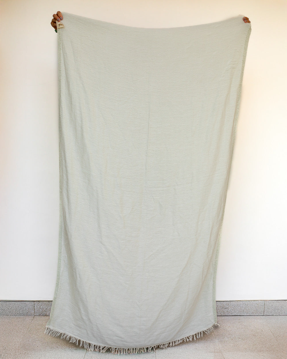 Eine Person hält das große, hellbeige Strandtuch Zen von Main Design aus OEKO-TEX-zertifizierter Baumwolle mit ausgefransten Rändern vor einer schlichten, cremefarbenen Wand und einem gefliesten Boden hoch und zeigt nur ihre Hände.