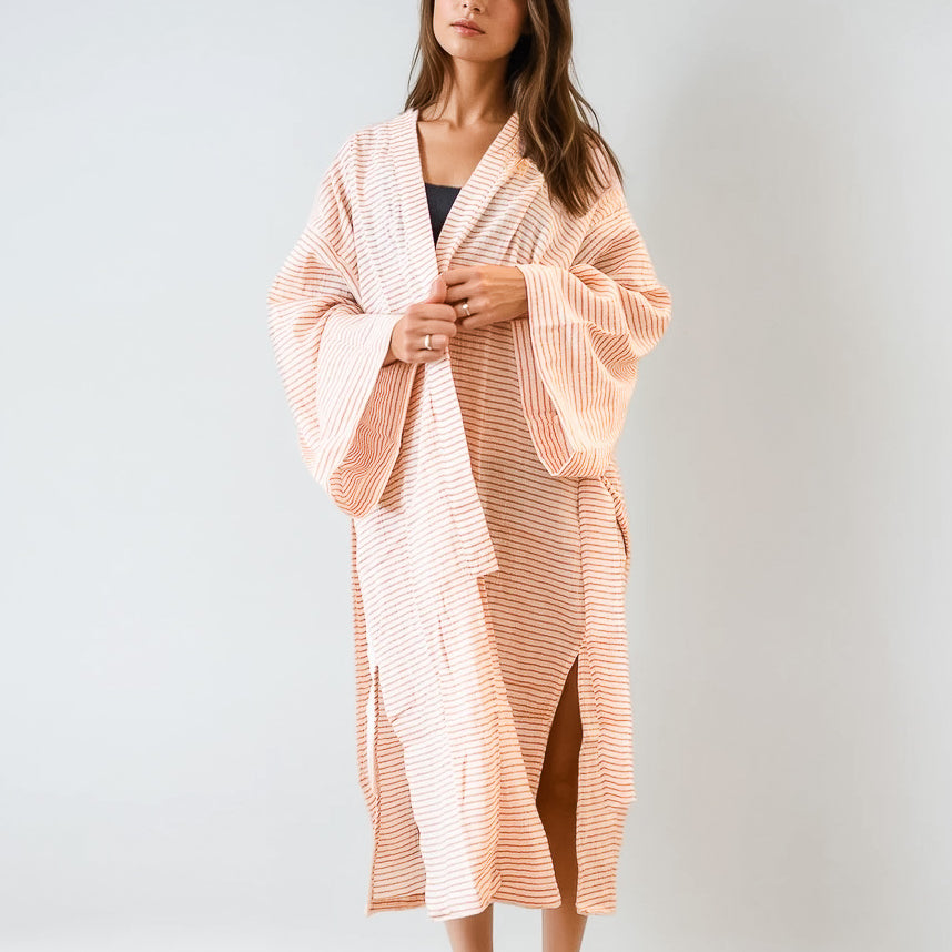 Eine Frau steht vor einem unifarbenen Hintergrund und trägt den Sunset Stripes Kimono von Main Design, ein langes, lockeres pfirsich-weiß gestreiftes Gewand mit weiten Ärmeln aus gekämmter Baumwolle, das über ein dunkles Oberteil für entspannte Strandmode gelegt wird.
