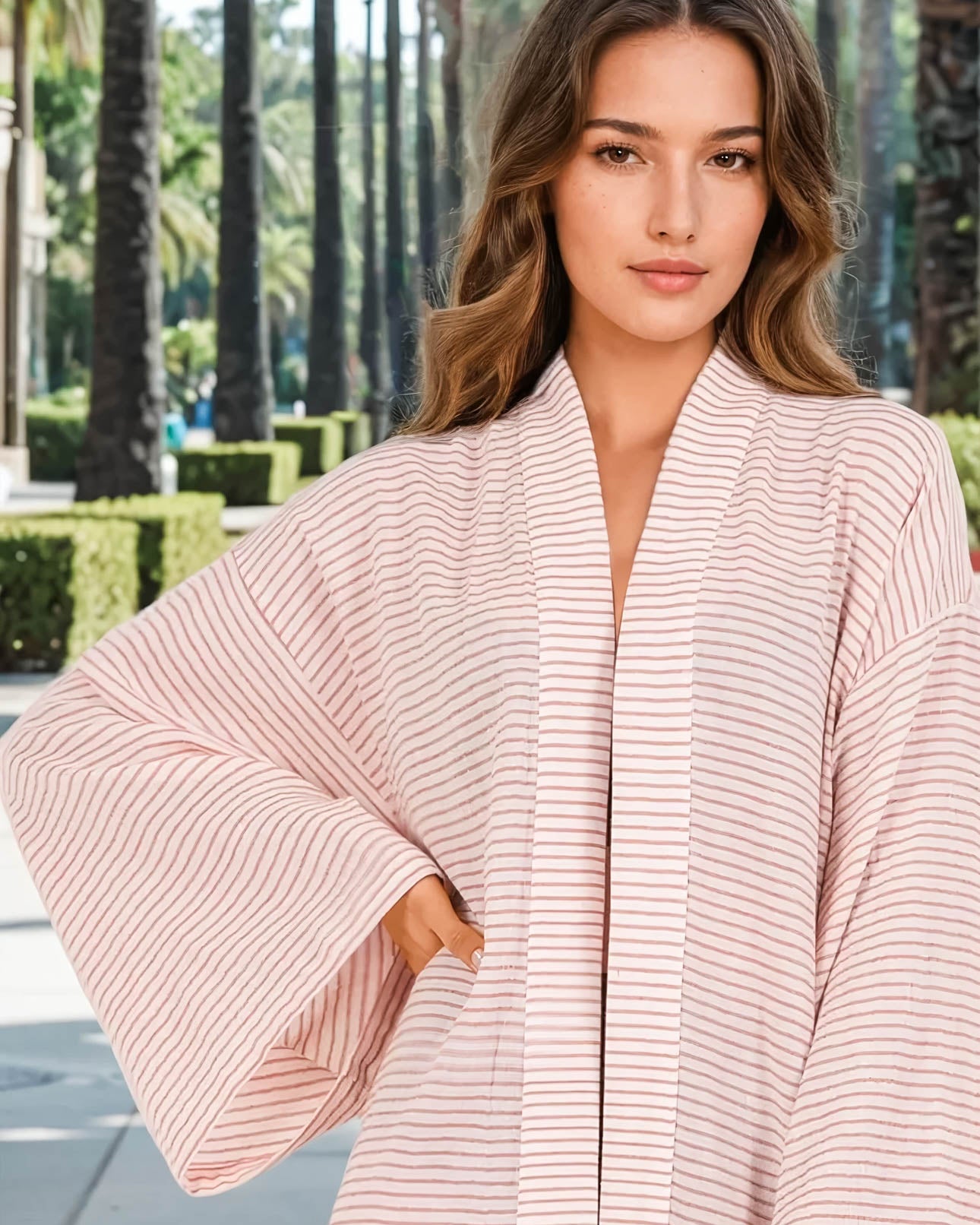 Eine Frau im Sunset Stripes Kimono von Main Design, mit weiten Ärmeln und einem hellen Rosaton, steht im Freien zwischen Palmen und gestutzten Büschen - ideal für einen entspannten Strandstil.