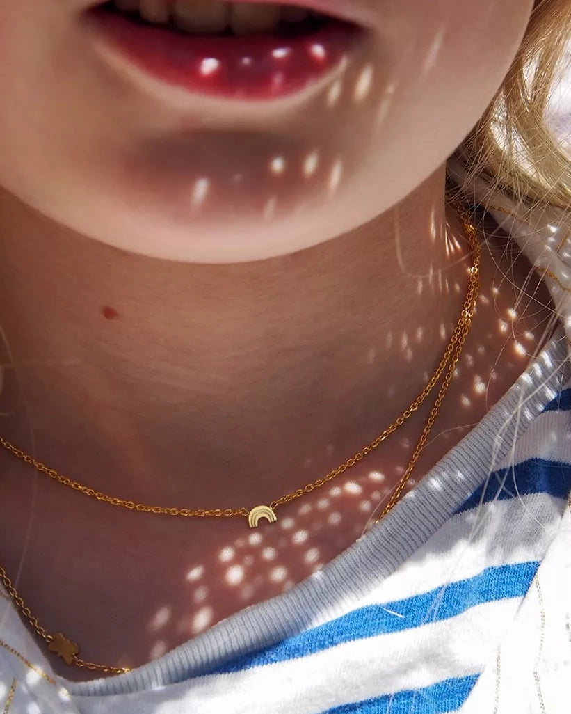 Nahaufnahme des unteren Gesichts und des Halses eines Kindes, das zwei 24K vergoldete Halsketten von Adorabili trägt, eine davon mit einem kleinen Mondsichelanhänger. Das Licht spielt auf der Haut und dem gestreiften Hemd - ein schönes Geschenk für Kinder von Adorabili.