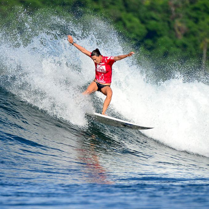 Ein Surfer in einem roten Trikot reitet mit ausgestreckten Armen auf einer Welle, umgeben von plätscherndem Wasser, während im Hintergrund ein üppiger grüner Wald verschwimmt.