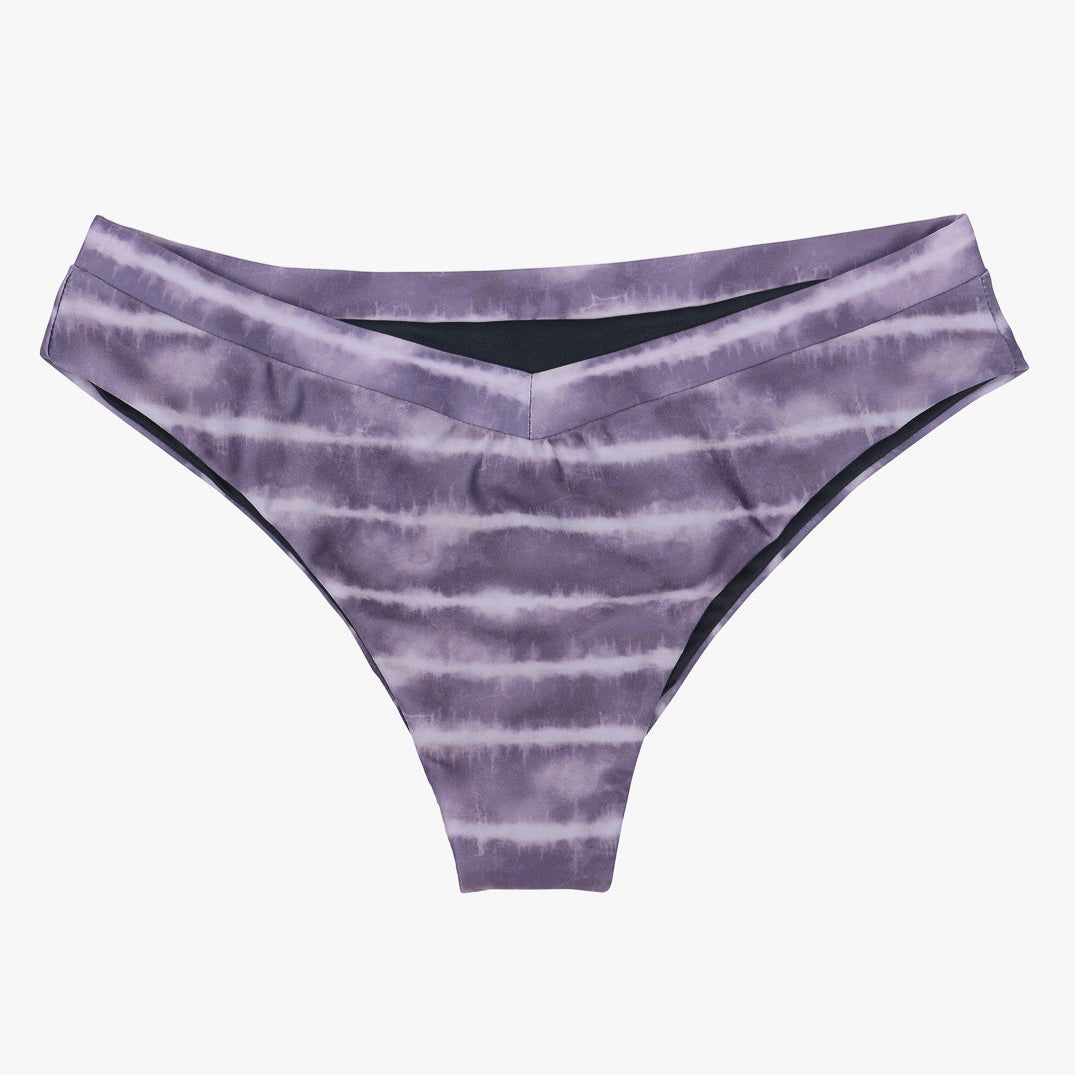 Ein Paar Main Design Ember Bikinihosen in lila Batikmuster. Der Stoff ist an den Seiten leicht gerafft, was ihm einen strukturierten Look verleiht. Diese tief sitzenden Bikinihosen im V-Schnitt sind aus recycelten Materialien gefertigt und auf einem schlichten weißen Hintergrund abgebildet.