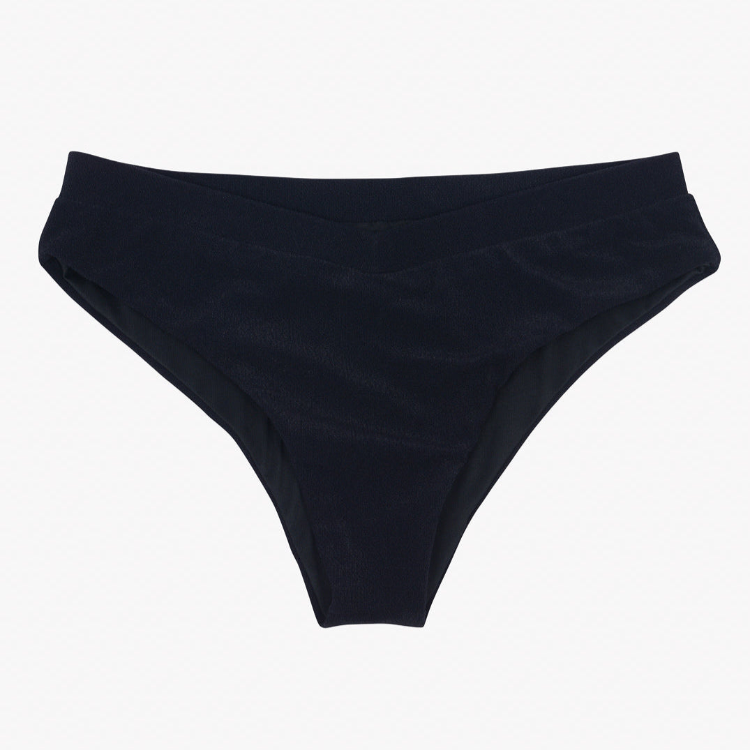 Ein schwarzes Bikinihöschen ist vor einem schlichten weißen Hintergrund zu sehen. Die Ember Bikinihose von Main Design hat einen leicht gebogenen Bund und ein V-Schnitt-Design und besteht aus einem scheinbar glatten, dehnbaren Stoff aus recycelten Materialien.