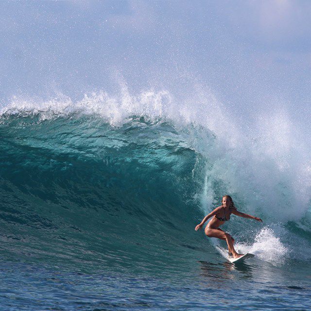 Eine Frau surft auf einer großen, blauen Meereswelle unter klarem Himmel und balanciert auf ihrem Surfbrett, während das Wasser hinter ihr plätschert.