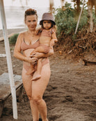 Eine Frau in einer beigen Main Design Gem Bikinihose hält ein kleines Kind mit Hut und passendem Badeanzug. Sie stehen an einem Sandstrand mit viel Grün im Hintergrund, im Schatten eines Strohdachs, während sich in der Ferne das Meer erstreckt.