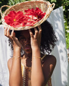 Eine Person mit lockigem Haar, die einen Weidenkorb mit großen roten Blumen auf dem Kopf trägt, steht im Freien im Sonnenlicht, verdeckt teilweise ihr Gesicht mit den Händen und trägt das Armband von Adorabili der Marke Adorabili.
