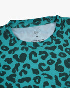 Eine Nahaufnahme des Aina Kinder UV-Shirts von Main Design in Blaugrün mit schwarzem Leopardenmuster. Ideal für sonnige Tage, bietet es LSF 50+ Schutz. Kragen und ein Teil der Brust sind sichtbar, ebenso wie ein weißes Etikett mit der Größe „S/M“ und der schnelltrocknenden Funktion.