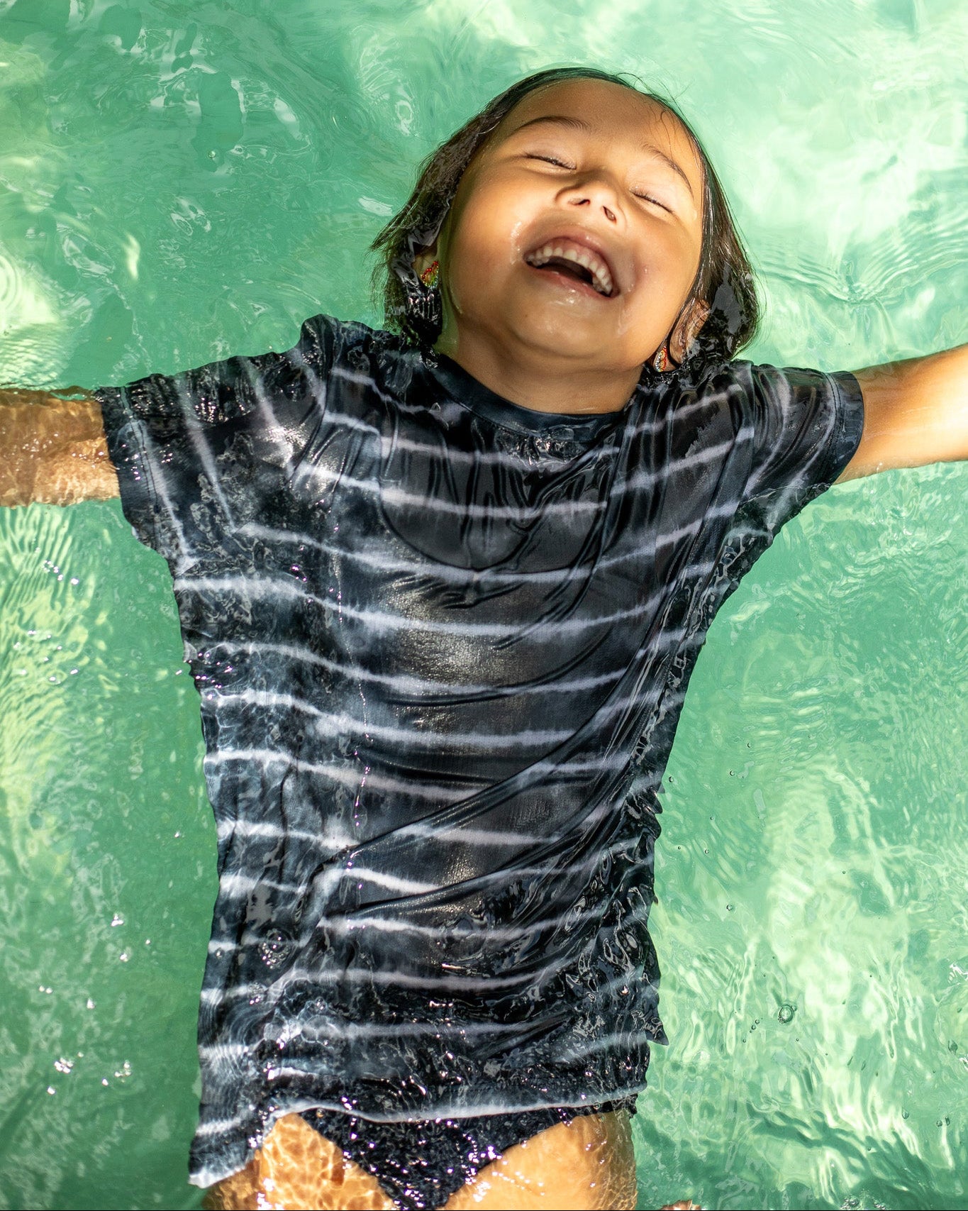 Ein Kind in einem dunkel gestreiften Aina Kinder UV-Shirt von Main Design schwimmt glücklich auf dem Rücken in einem klaren, sonnenbeschienenen, grünlichen Pool. Das Shirt aus nachhaltigem Stoff bietet UPF 50+ Sonnenschutz für sicheres Spielen.