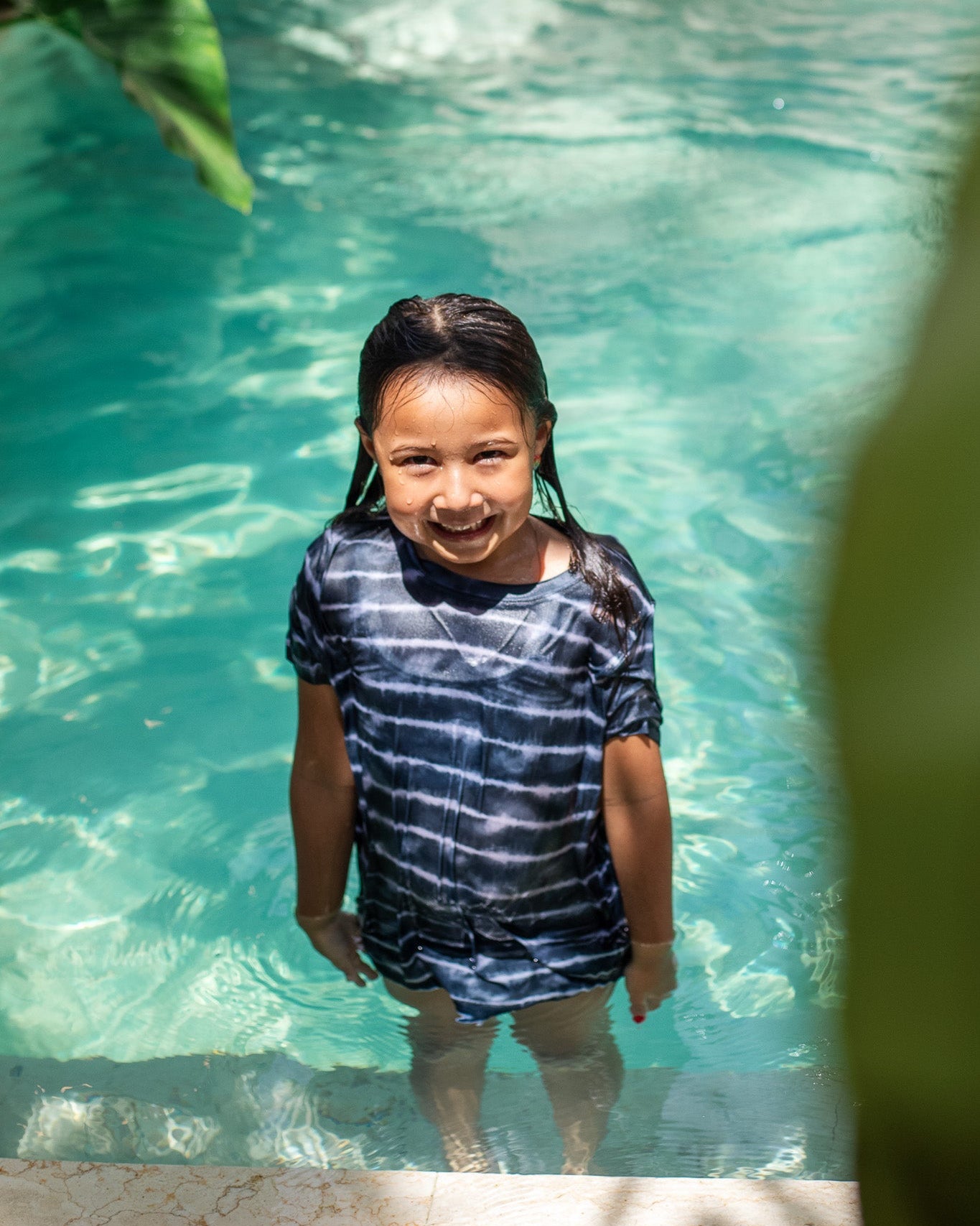 In einem klaren, türkisfarbenen Pool trägt ein Kind mit nassen Haaren und einem Lächeln ein dunkles Aina Kinder UV-Shirt von Main Design mit Batikmuster. Das Sonnenlicht spiegelt sich im Wasser und verleiht der fröhlichen Szene Helligkeit. Mit UPF 50+-Schutz und grünen Blättern im Hintergrund ist es ein idealer sonniger Tag.