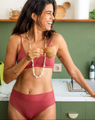 Eine Frau mit langen dunklen Haaren, die eine rote Alice Bikinihose von Main Design und eine weiße Perlenkette trägt, steht in einer Küche. Sie lächelt und blickt nach rechts, während sie in ihrer linken Hand eine kleine Tasse hält. Die Küche hat grüne Schränke, Holzregale und weiße Wände.