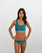 Eine Frau mit langen dunklen Haaren steht vor einem hellen Hintergrund und trägt die Alice Bikinihose von Main Design. Die türkisfarbene Bikinihose hat ein dunkles Leopardenmuster und ist aus nachhaltigen Materialien gefertigt. Sie blickt mit den Händen in die Hüften in die Kamera.