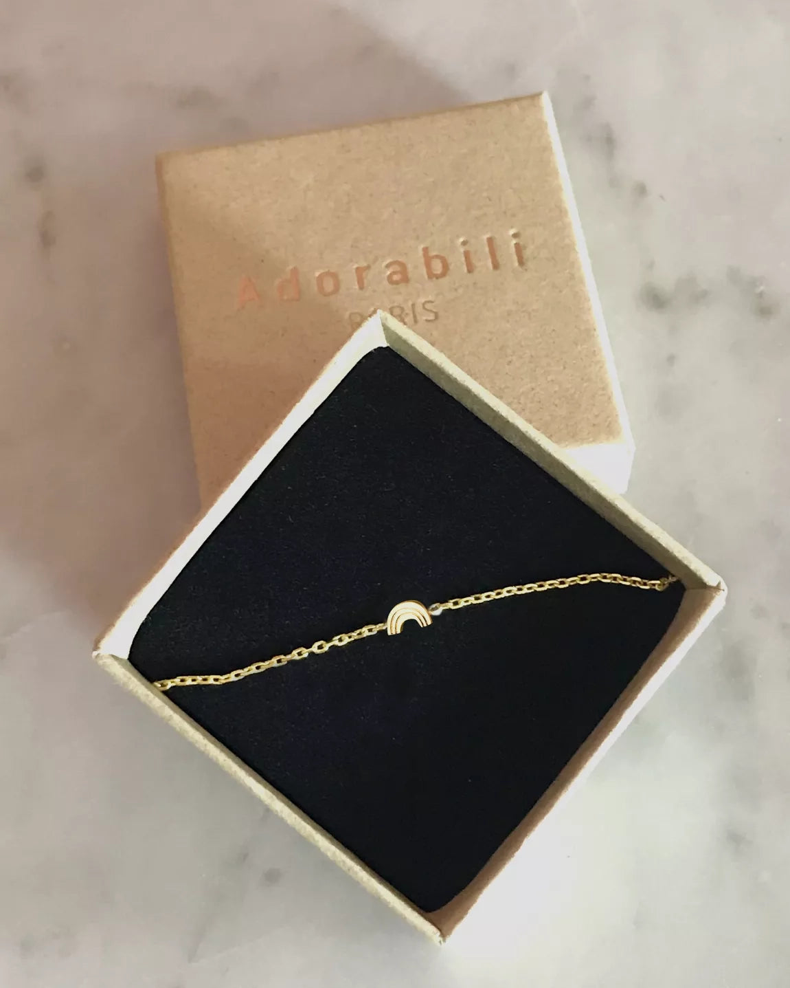 Das Armband von Adorabili, handgefertigt und 24K vergoldet mit Regenbogenanhänger, liegt in einer schwarzen Samtschachtel. Der braune Kartondeckel mit "Adorabili Paris" steht auf einer Marmorfläche dahinter.
